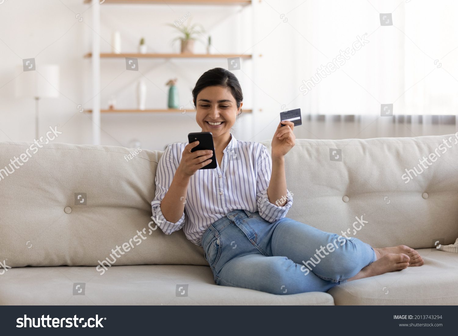 Excited Indian woman paying online by credit card for goods, using smartphone, sitting on couch at home, happy young female satisfied customer browsing internet banking service on device, shopping #2013743294