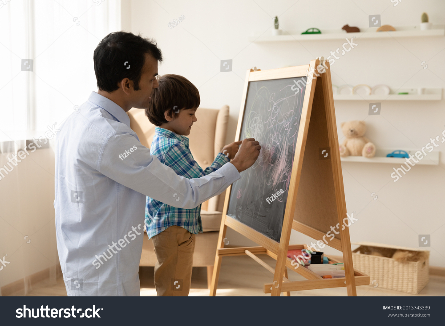 Loving Indian father with little son drawing on black board with colored chalks together, painting, young dad and preschool boy child having fun at home, family involved in creative activity #2013743339