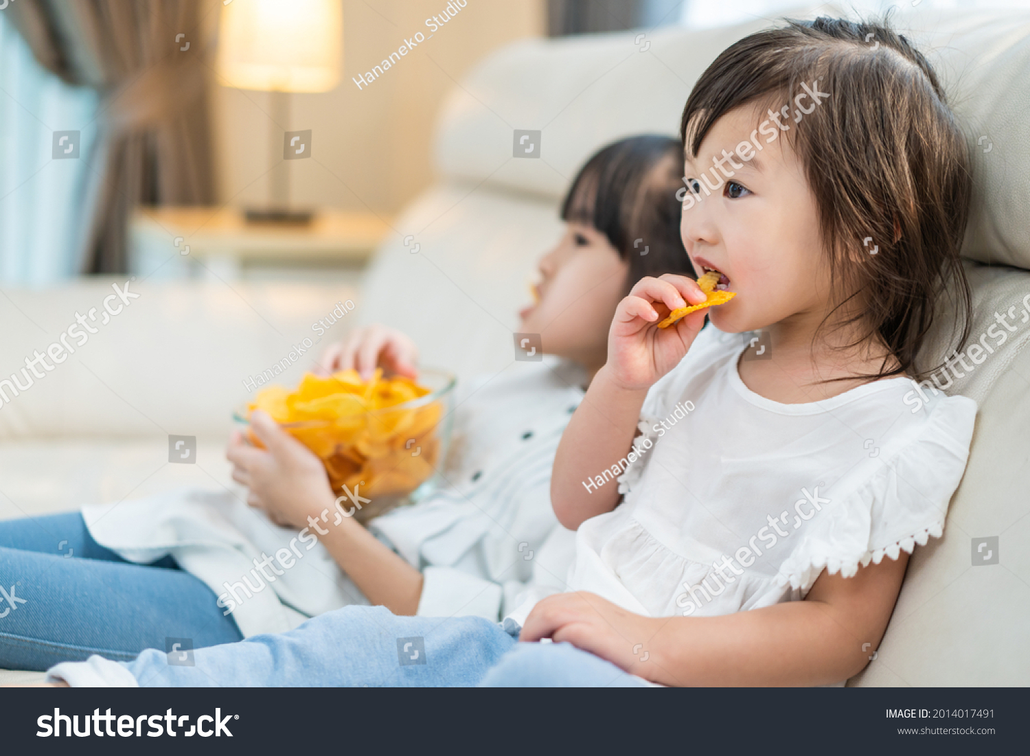Asian Hungry little girl sibling sisters Puts snack in mouth with hand. Two sweet preschool kid resting on sofa at home enjoy eat fast food, potato chips. Tasty and unhealthy food for children concept #2014017491