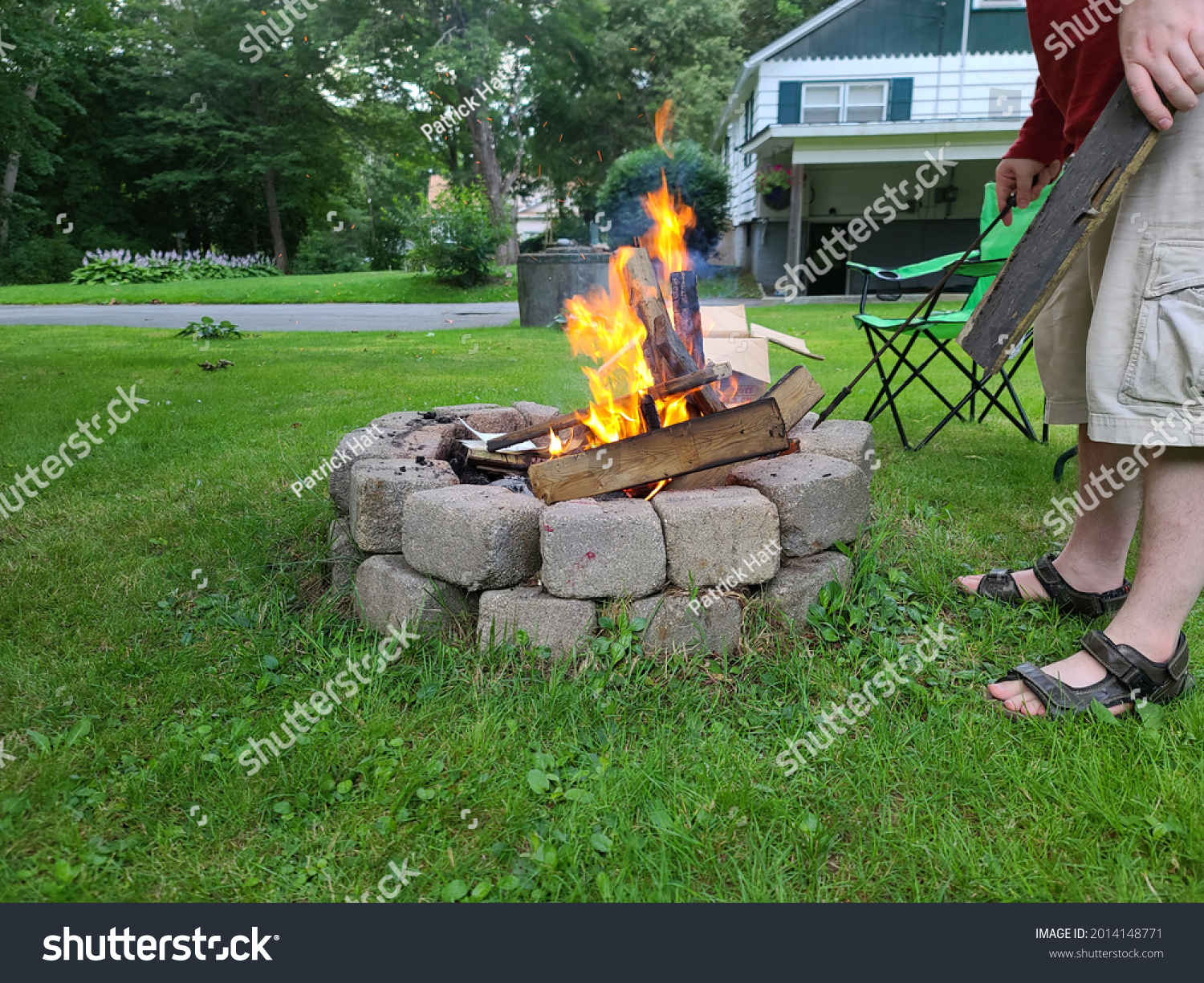 The view of a backyard fire pit. The The fire is lit and is burning shredded paper while igniting boards in the round, cinder block fireplace. #2014148771