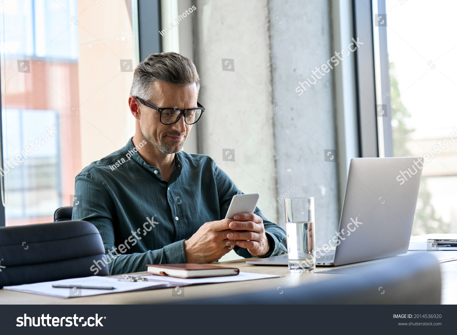Smiling mature businessman holding smartphone sitting in office. Middle aged manager ceo using cell phone mobile apps and laptop. Digital technology applications and solutions for business development #2014536920