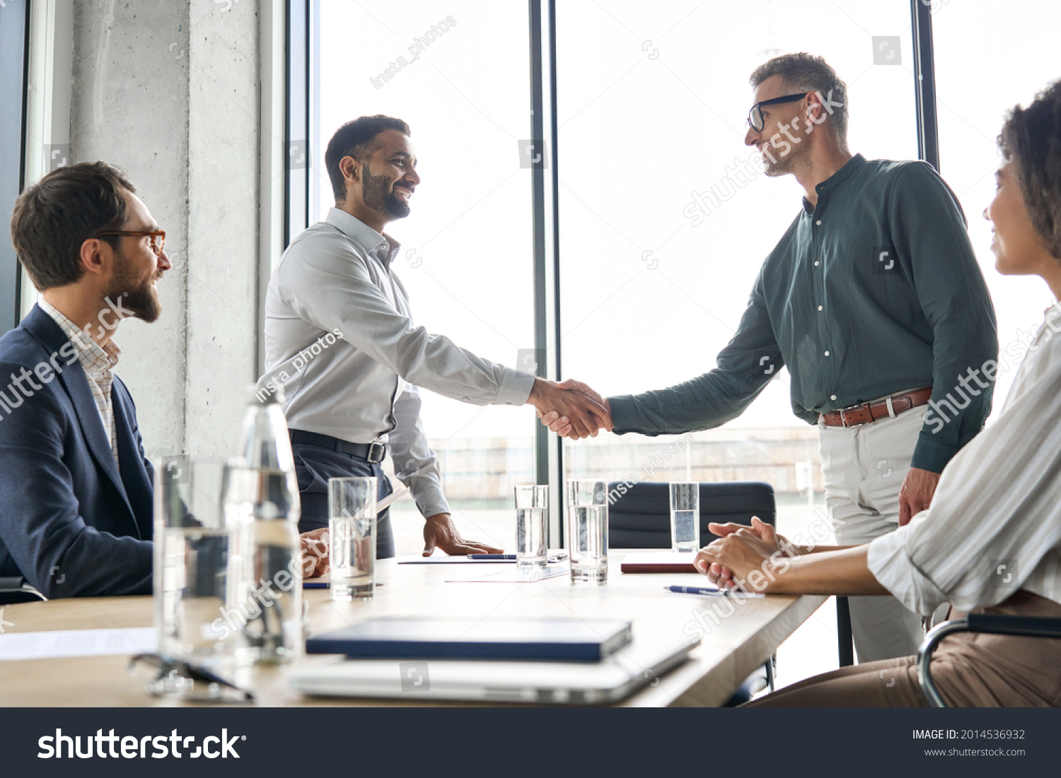 Two happy diverse professional business men executive leaders shaking hands after successful financial deal at group board office meeting. Trust agreement company trade partnership handshake concept. #2014536932