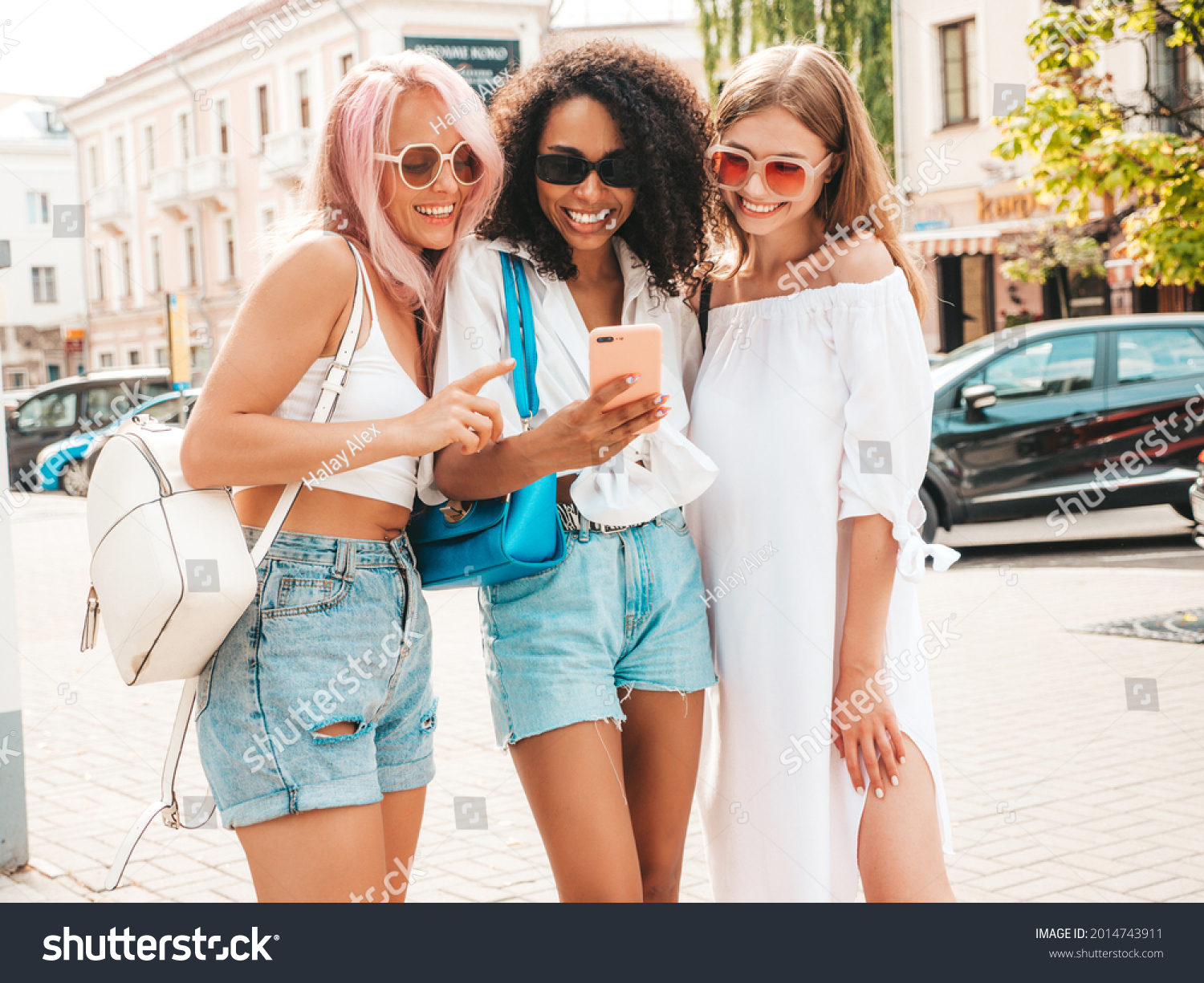 Three young beautiful smiling hipster female in trendy summer clothes.Sexy carefree multiracial women posing in the street.Positive models in sunglasses. Looking at smartphone screen.Using phone apps #2014743911