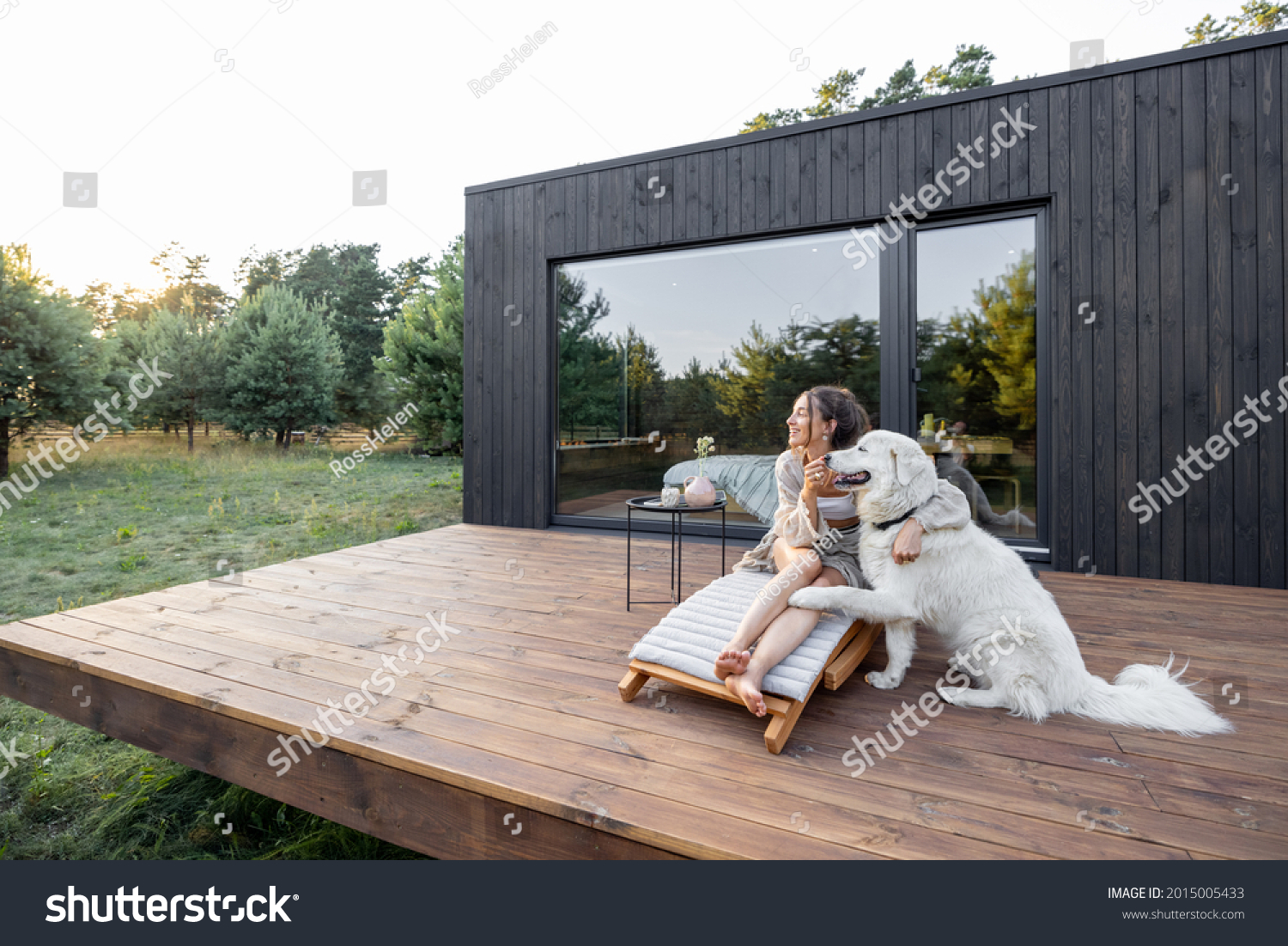 Woman enjoys the nature while sits on sunbed on wooden terrace near the modern house with panoramic windows near pine forest while hugs her pet. Concept of solitude and recreation on nature #2015005433