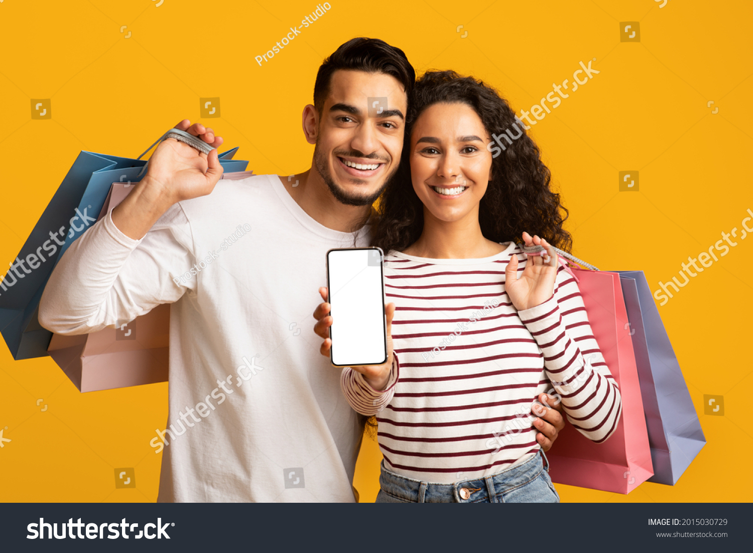 Online Shopping App. Cheerful young arab couple showing smartphone with blank white screen at camera and holding bright shopper bags while standing isolated over yellow studio background, mockup #2015030729