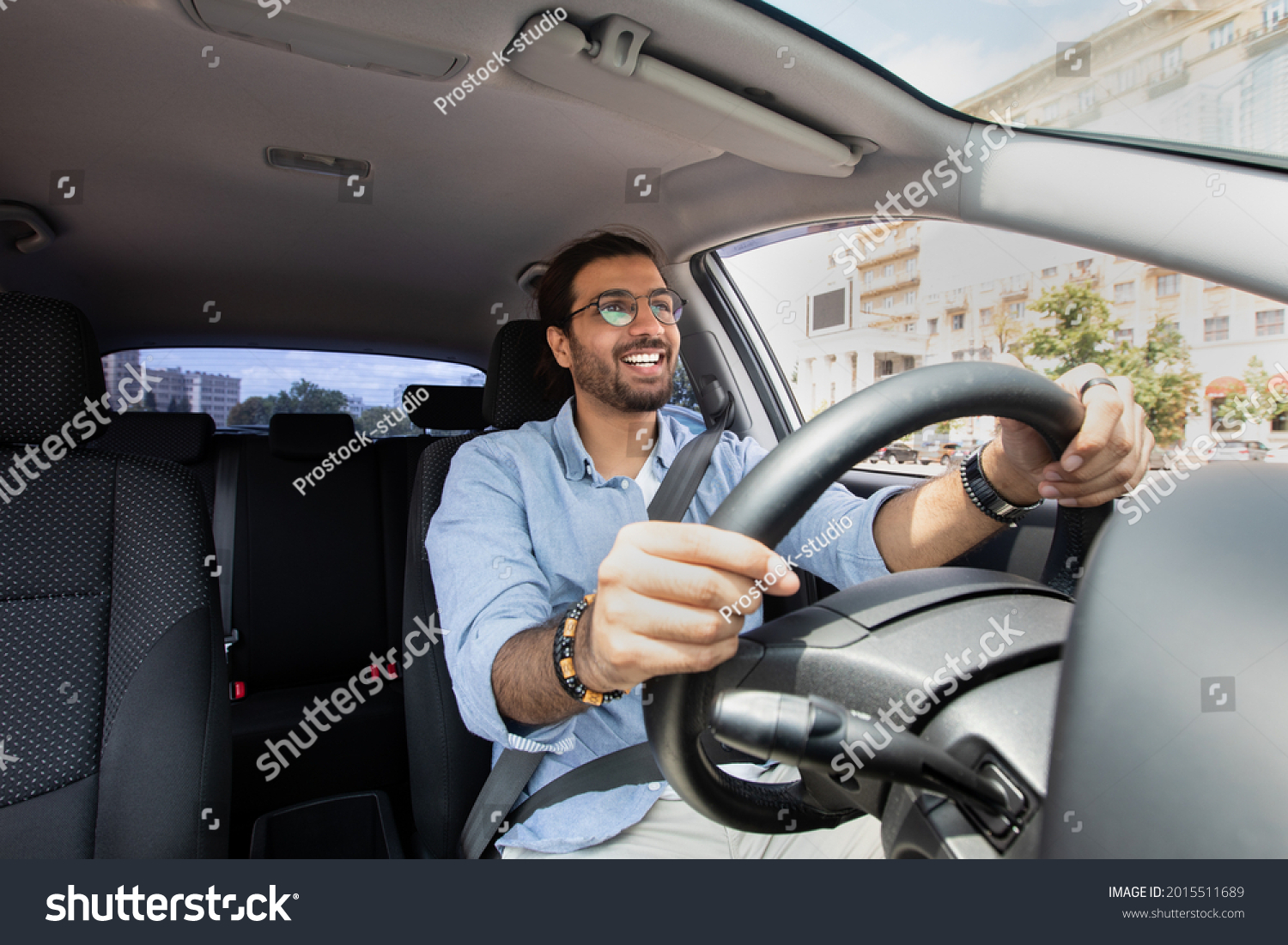 Joyful indian man driving car, shot from dashboard, going on trip during summer vacation, copy space. Happy middle-eastern guy in casual outfit and glasses driving his brand new nice car #2015511689