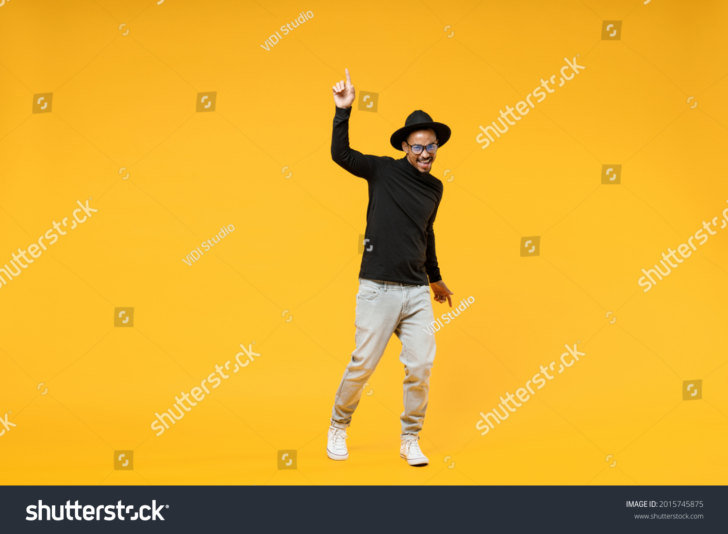 Full length young fun african man 20s wearing stylish black hat shirt eyeglasses doing winner gesture pointing index finger up walk going isolated on yellow orange color background studio portrait. #2015745875