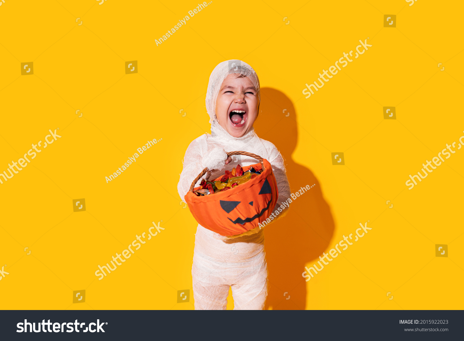Child in mummy costume holding basket of chocolates in front of yellow background. #2015922023