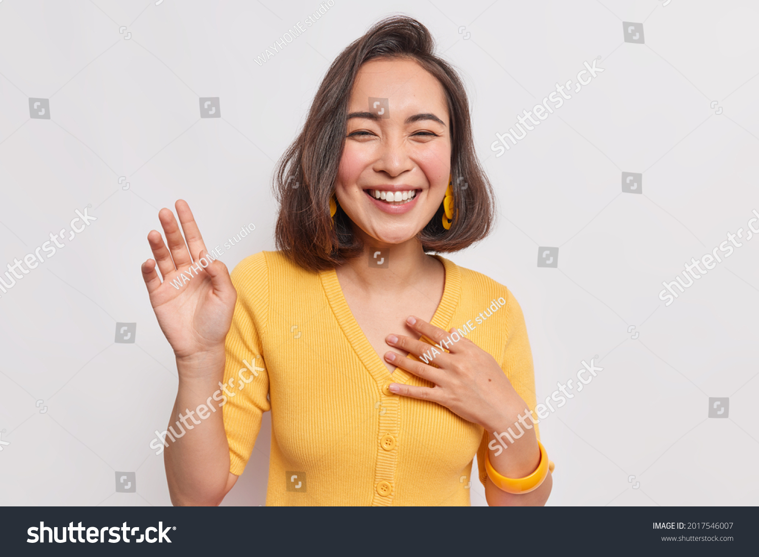 Happy dark haired pretty Asian woman with positive expression laughs joyfully keeps hand raised smiles broadly wears yellow jumper earrings hears something funny isolated over white background. #2017546007