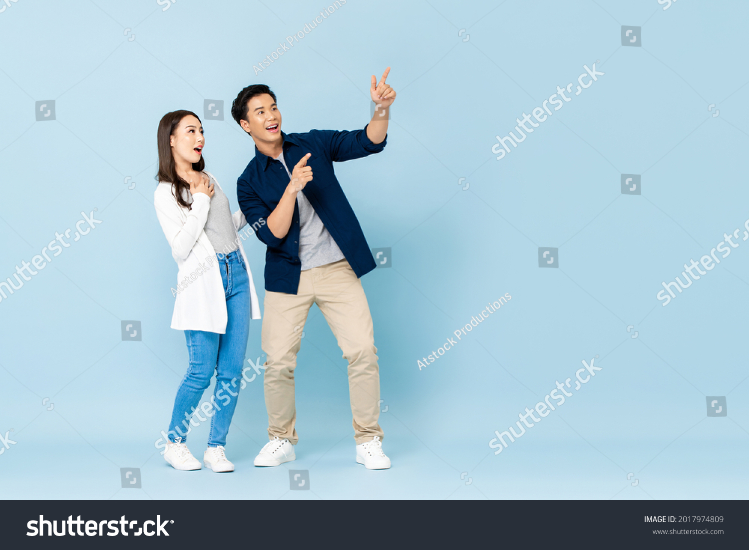 Full length portrait of happy excited Asian couple tourists pointing hands to empty space on isolated light blue background #2017974809