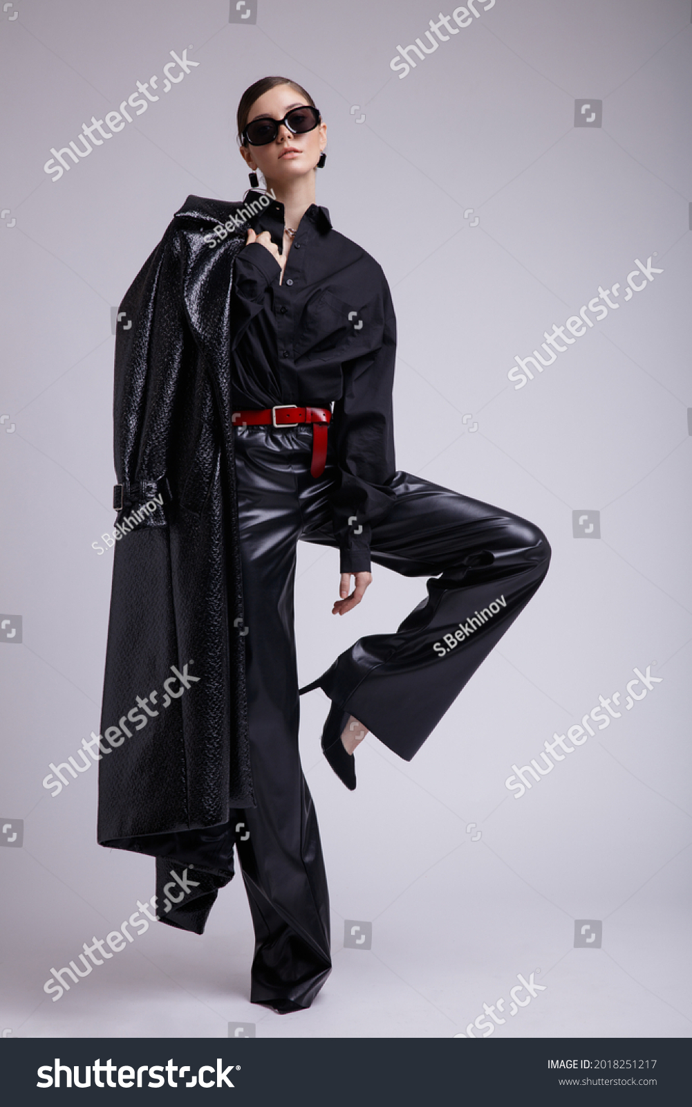 High fashion photo of a beautiful elegant young woman in a pretty leather coat, pants, stylish sunglasses, accessories, shirt, red belt posing over gray background. Studio Shot. Gathered dark hair #2018251217