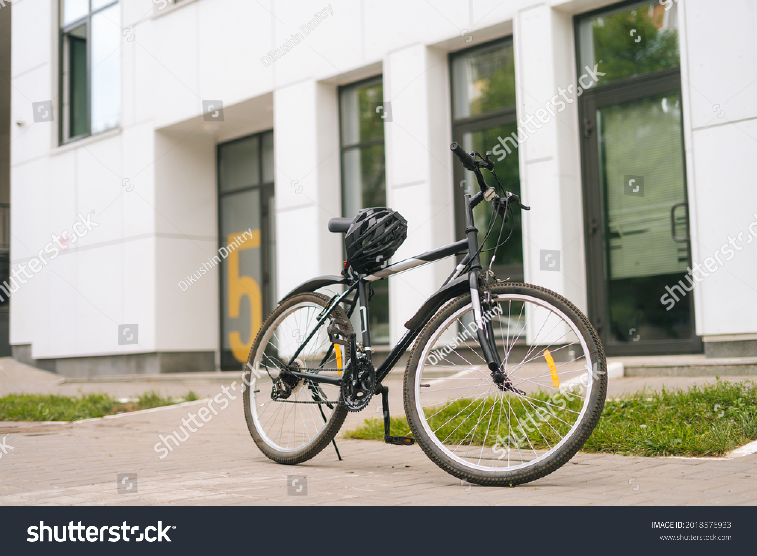 Modern bicycle standin on footplate in city street on blurred background of front door of apartment building, office building. Protective helmet lies on seat of bike, outdoors, no people, nobody. #2018576933