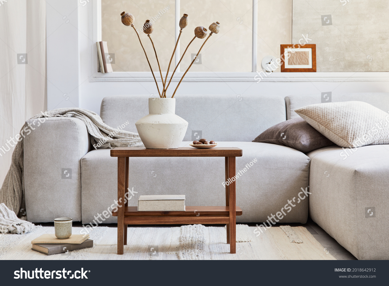 Creative composition of stylish living room interior with grey corner sofa, window, wooden coffee table, vase with dried flowers and personal accessories. Beige neutral colors. Template. #2018642912