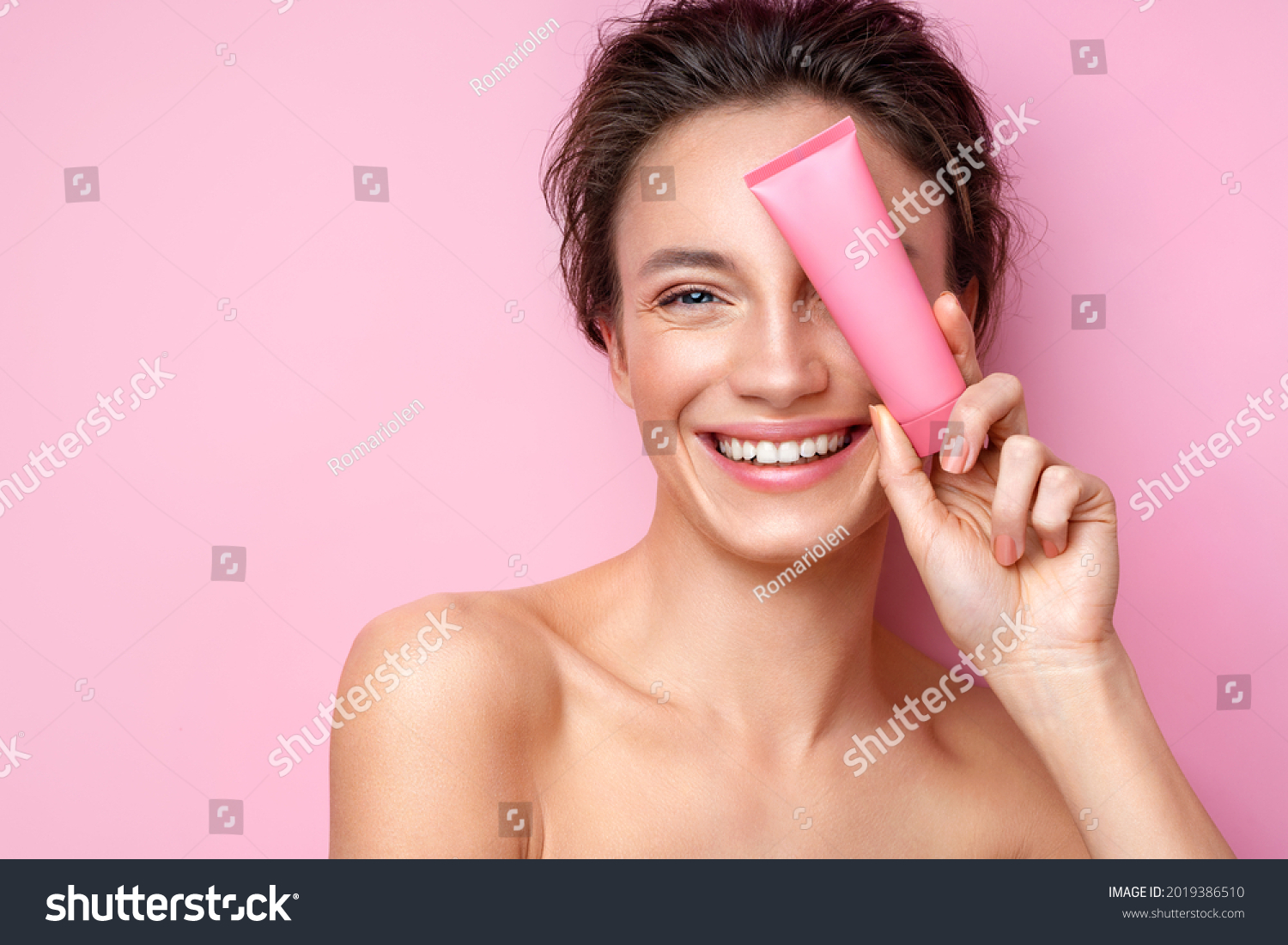 Smiling woman holds tube with cosmetic cream. Photo of attractive woman with perfect makeup on pink background. Beauty concept #2019386510