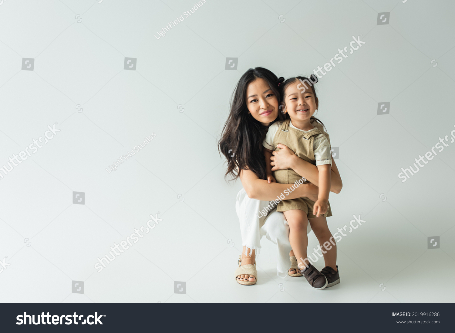 full length of stylish asian mother hugging toddler daughter on grey #2019916286