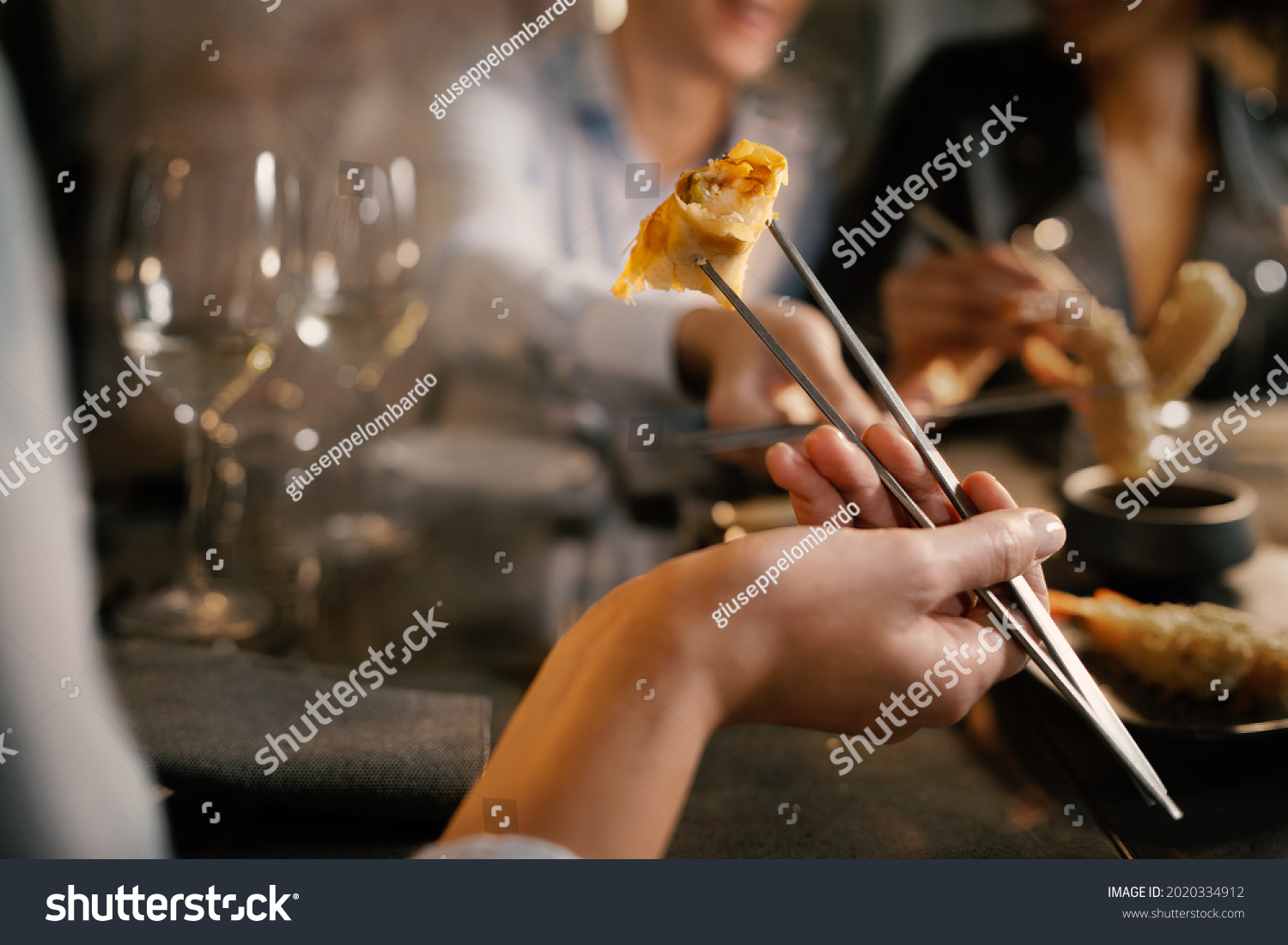 Detail of a woman's hand grabbing a spring roll with metallic chopsticks. He sits at a table with friends in a fancy asiatic fusion food restaurant. #2020334912