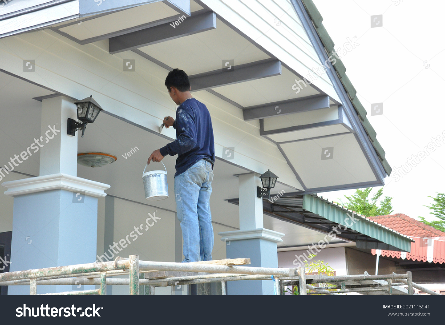 a painter one hand carry color bucket and other hand hold brush standing on scaffolding,painting house gable.worker man paint exterior house wall,people work at construction,maintenance or repair home #2021115941