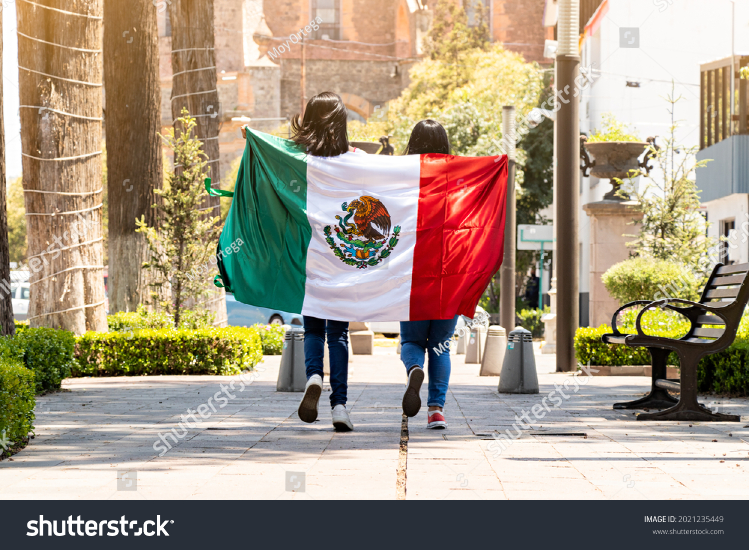 two young mexican women running on a walkway on a tree-lined street, with the mexican flag #2021235449