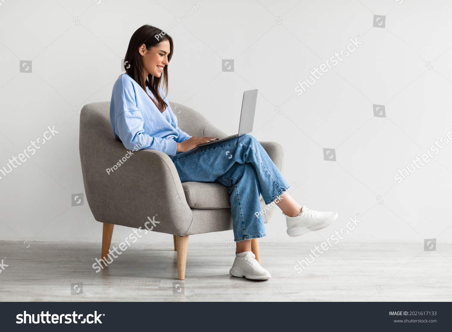 Smiling young woman in casual wear working online, sitting in armchair and using laptop against white studio wall, copy space. Cheerful Caucasian lady surfing internet on portable pc #2021617133