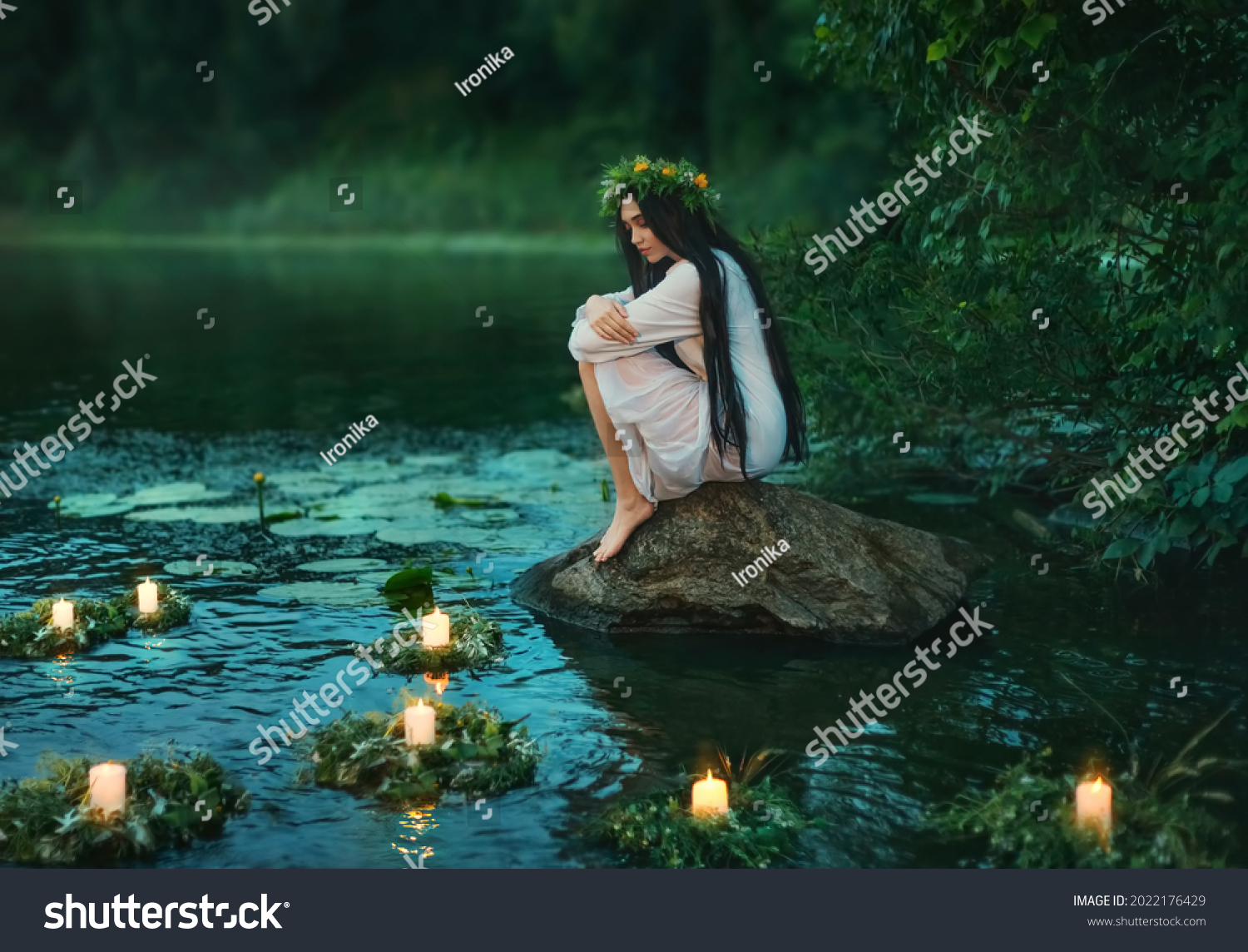 Slavic girl sits on stone on shore lake. Nymph fantasy woman hugs knees. Long black hair. Wreaths of grass, flowers float on water. Candles burning. River dusk forest green tree. Riutal of Divination. #2022176429
