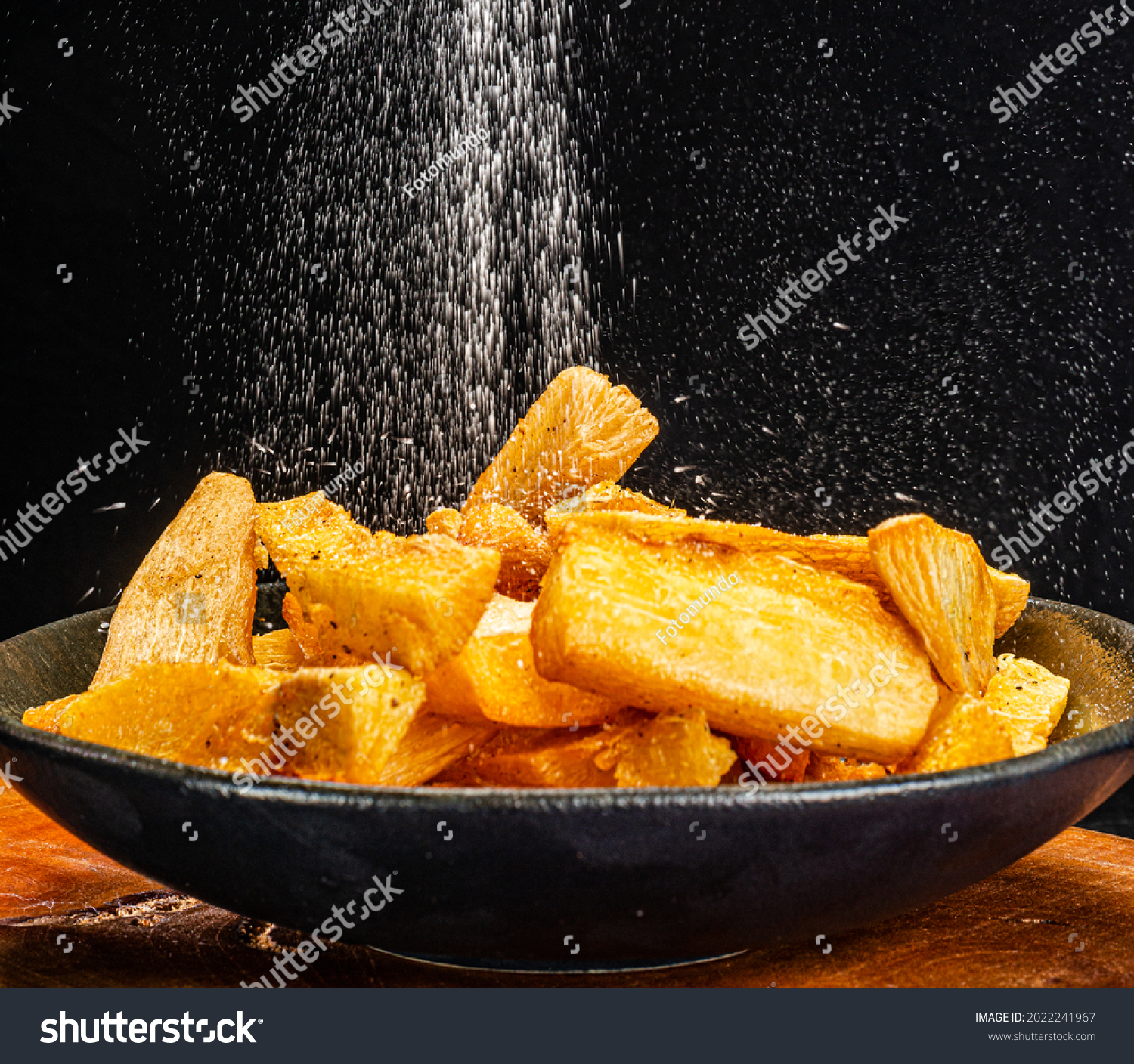 Salt being poured over Fried manioc portion, traditional Brazilian bar food, served in a rustic dish on wood and black background. selective focus #2022241967