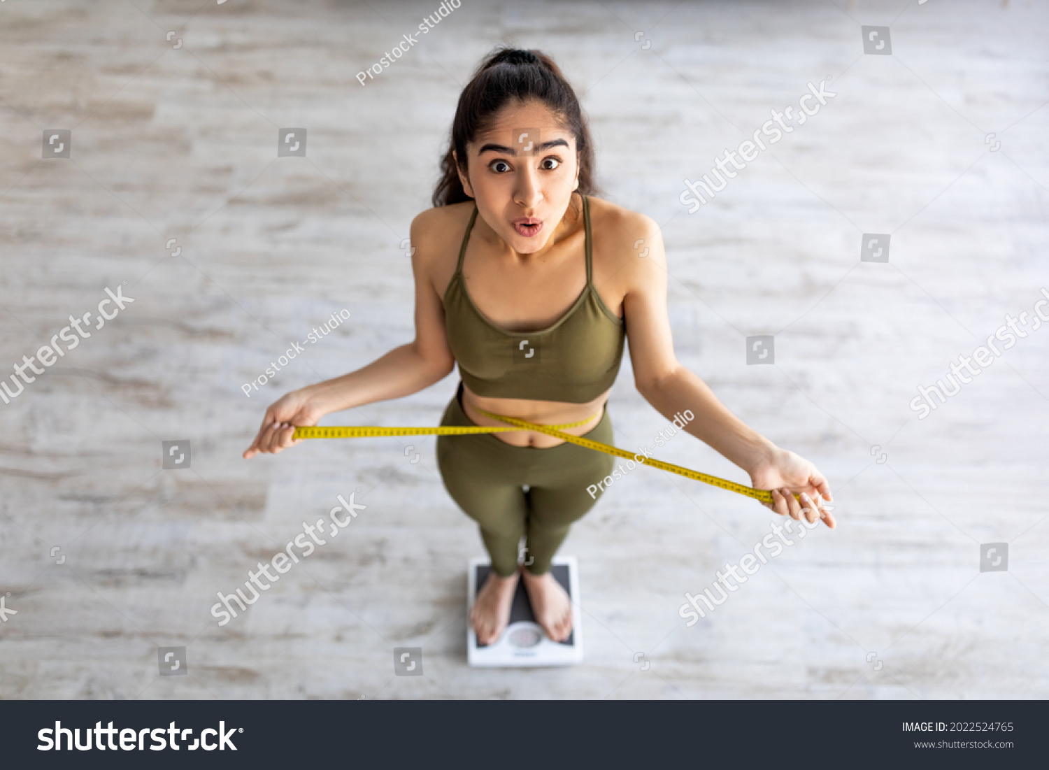 Above view of shocked Indian lady standing on scales, checking her waist measurements, surprised with unexpected results of weight loss program, indoors. Slimming diet and workout concept #2022524765