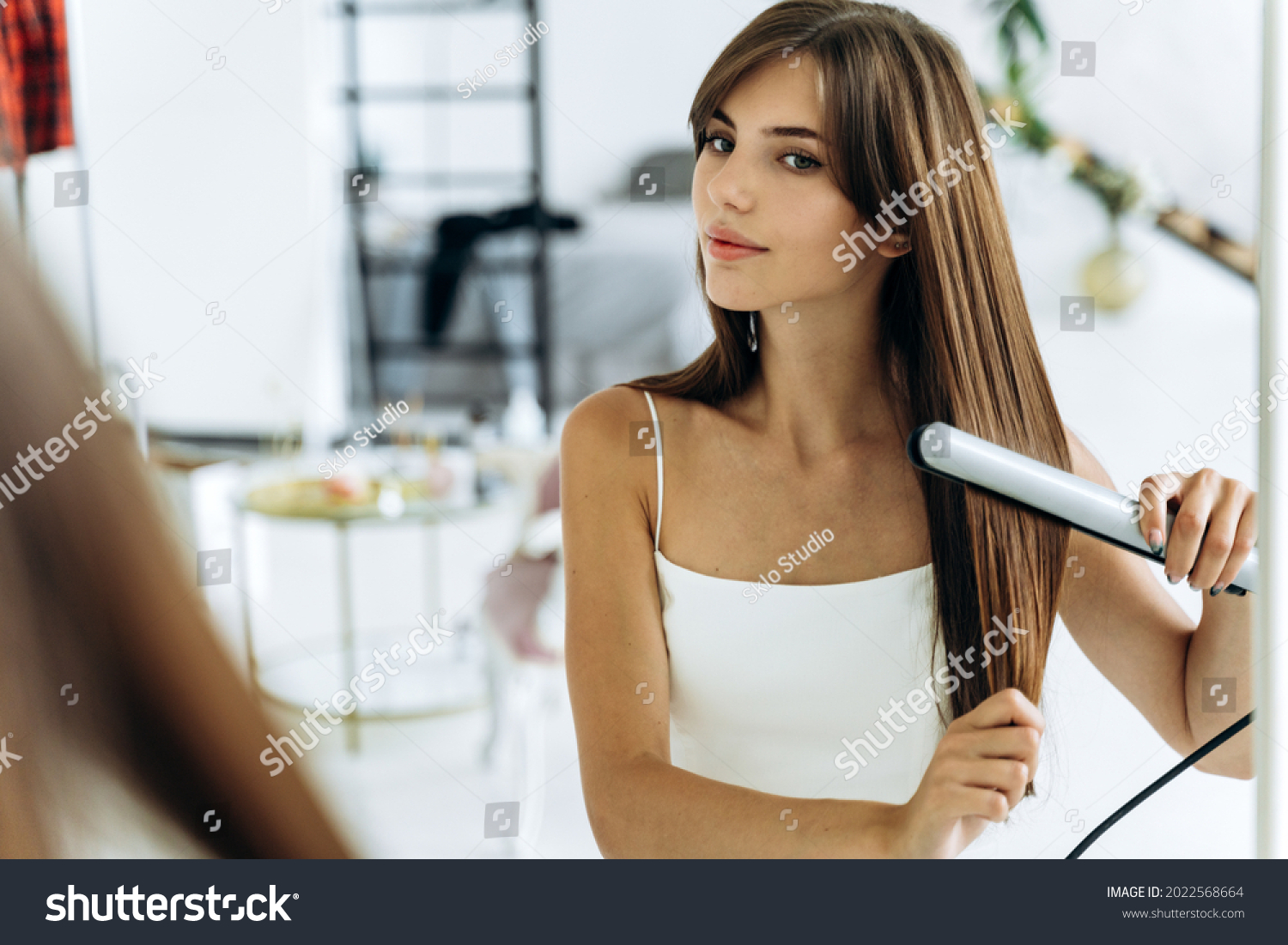 Beautiful young smiling woman using a hair straightener while looking into the mirror in bathroom. Confident girl looking at her reflection with pleasure smile  #2022568664
