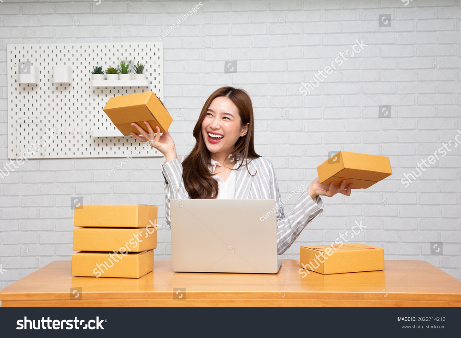 Happy young asian woman startup small business freelance holding parcel box and computer laptop and sitting on chair, Online marketing packing box delivery concept #2022714212