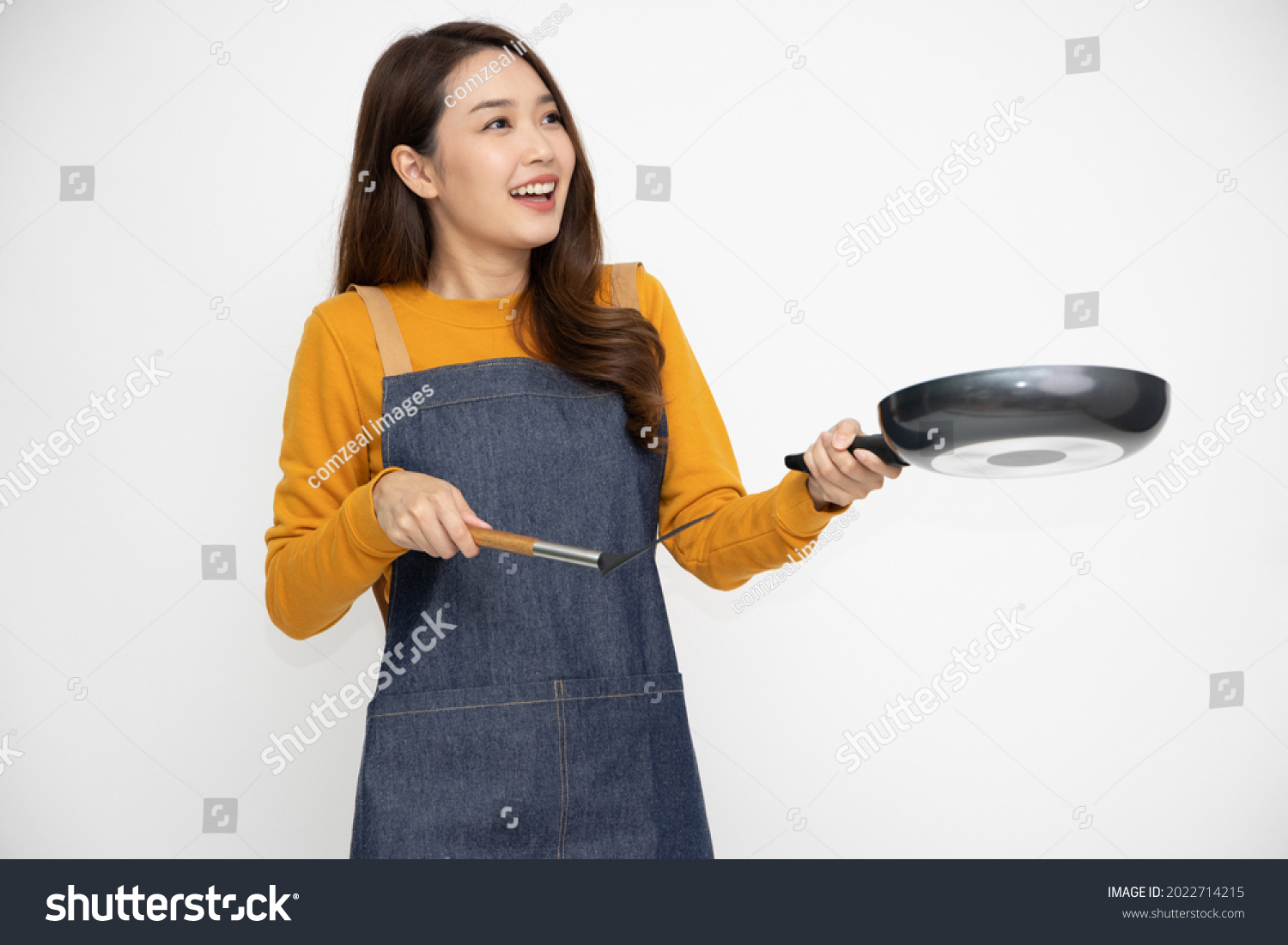 Happy young Asian woman housewife wearing kitchen apron cooking and holding pan and spatula isolated on white background #2022714215