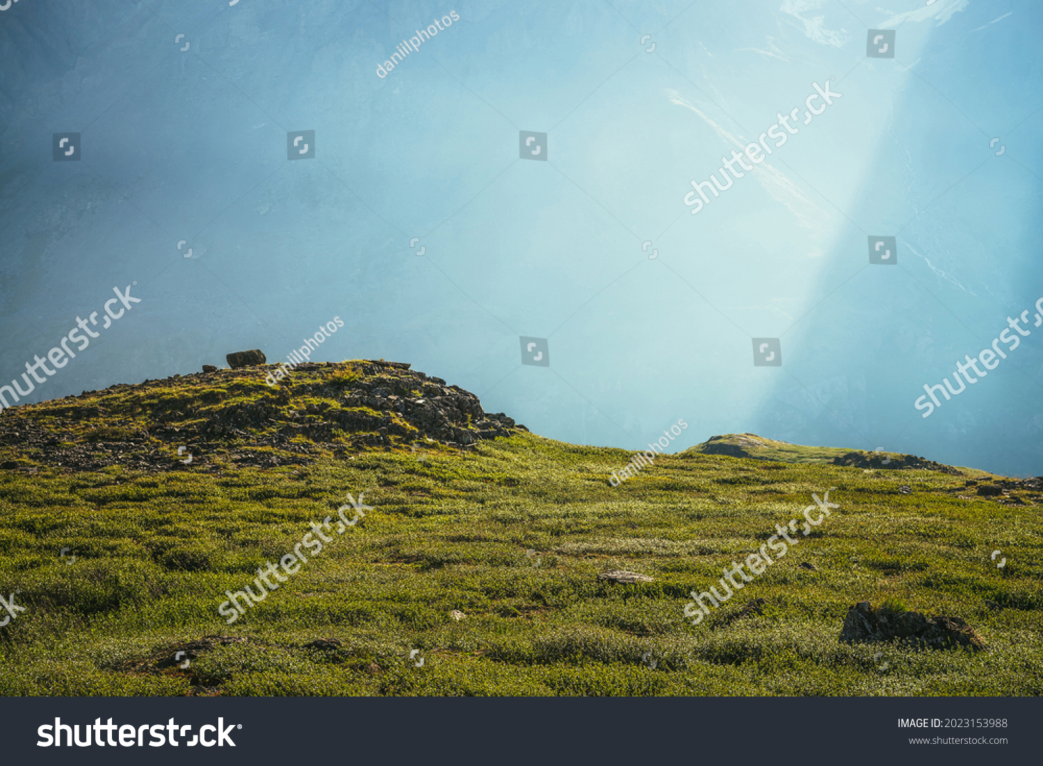Colorful green landscape with rocks and hills on background of giant mountain wall in sunlight. Minimalist vivid sunny scenery with sun beams and solar flare. Minimal alpine view. Scenic minimalism. #2023153988