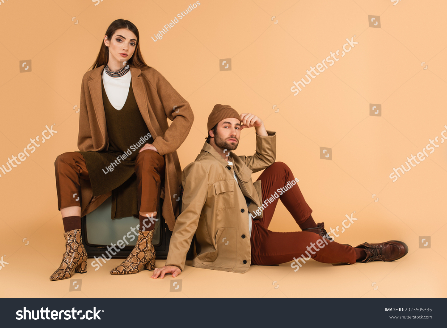 trendy woman sitting on vintage tv set near stylish man on floor on beige background #2023605335