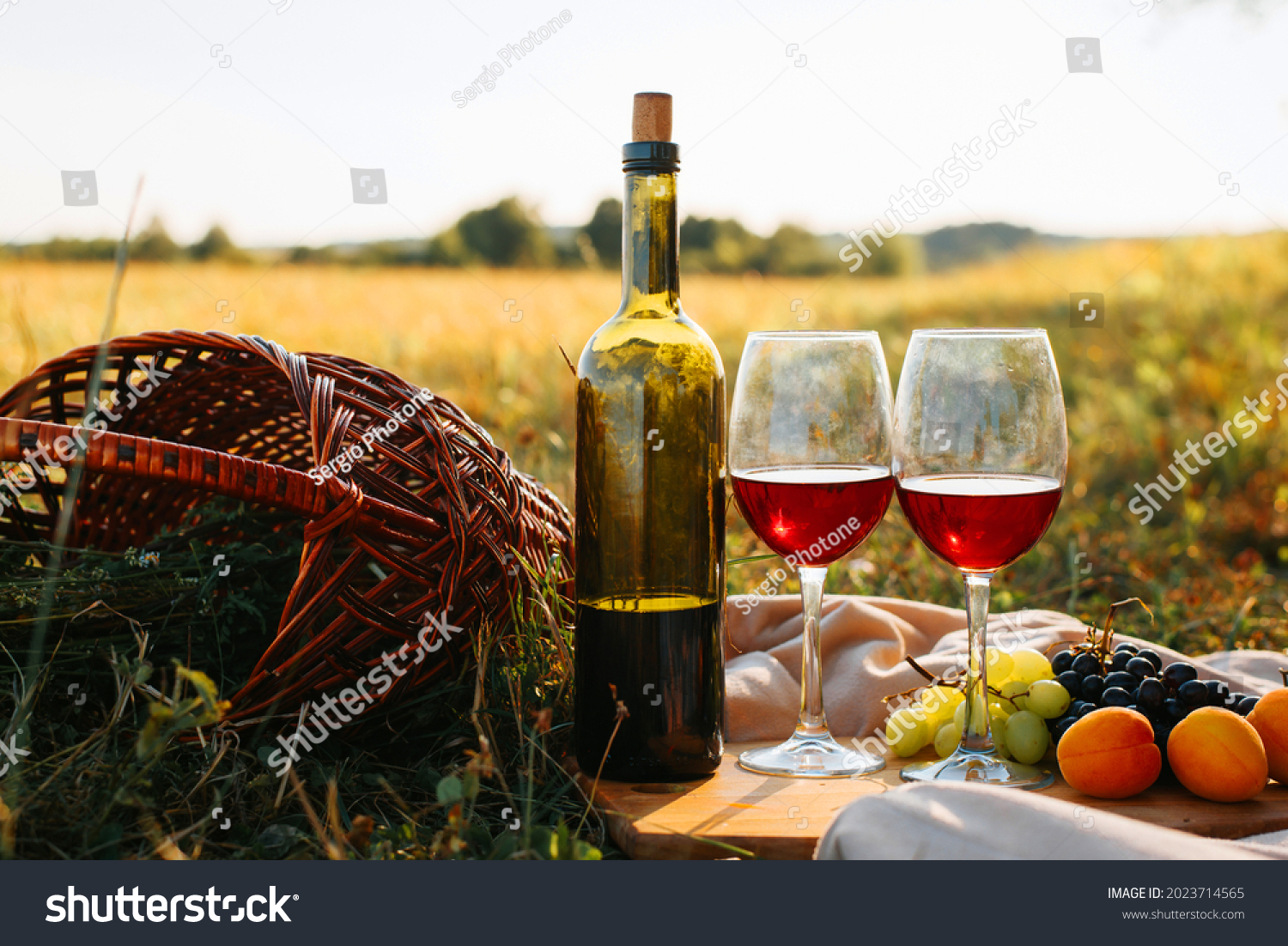 Picnic, romantic evening in nature concept. Bottle of red wine, glasses with drink, grapes, peaches on wooden board, wicker picnic basket on sunset background #2023714565