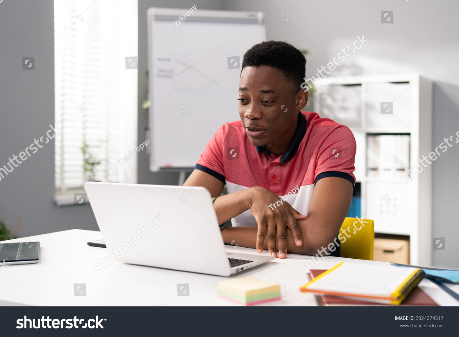 Trainee in office sits at desk in front of laptop monitor in background whiteboard, young dark-skinned man talks online with clients, co-workers, uses webcam, video call #2024274317