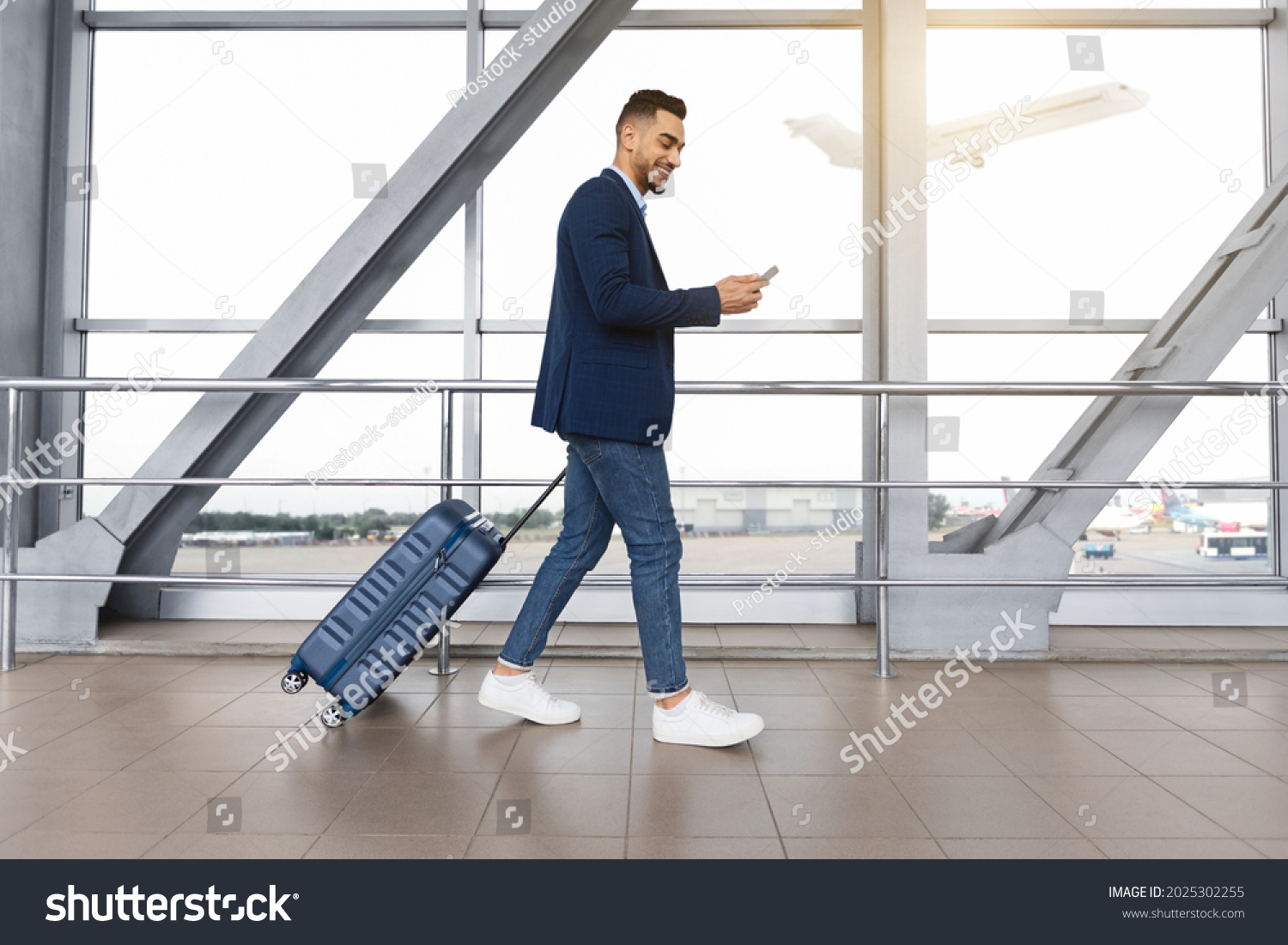 Handsome arab guy using smartphone while walking with suitcase at airport terminal, young middle eastern man browsing mobile internet on cellphone while going to flight boarding, side view #2025302255