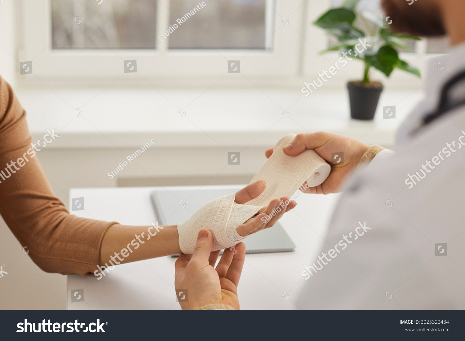 Doctor wrapping young woman's injured hand, close up. Doctor or nurse at modern medical office wrapping patient's sprained wrist with bandage. First aid and professional limb injury treatment concept #2025322484