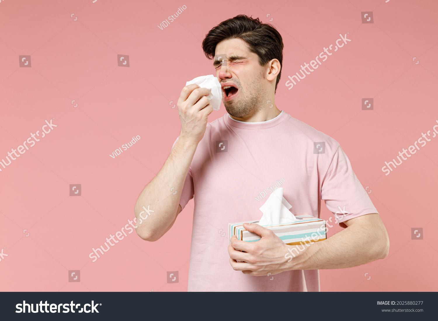 Sick unhealthy ill allergic man has red watery eyes runny stuffy sore nose suffer from allergy trigger symptoms hay fever hold paper napkin handkerchief isolated on pastel pink color background studio #2025880277