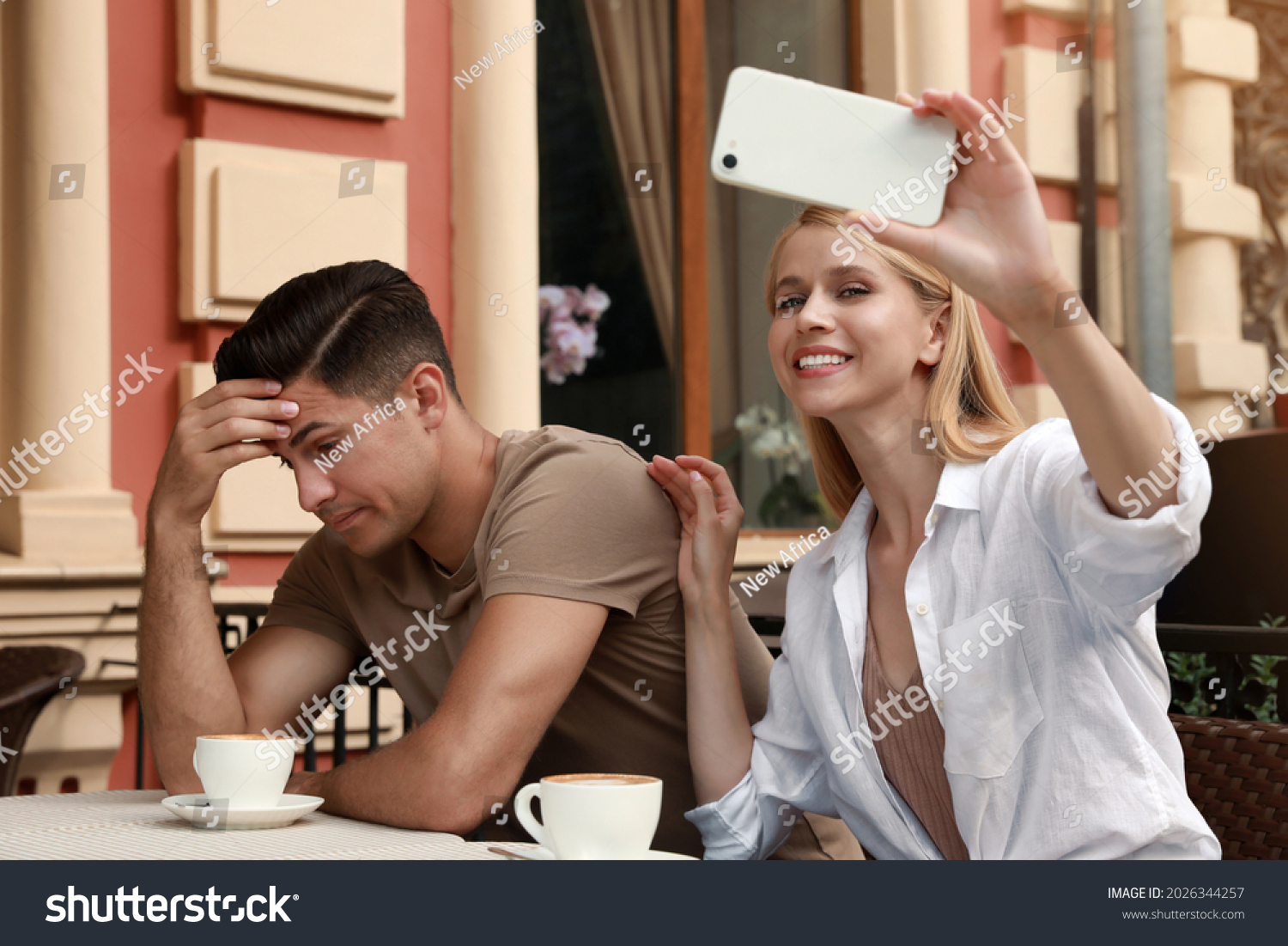 Young woman trying to take selfie with her displeased boyfriend in outdoor cafe. Boring date #2026344257