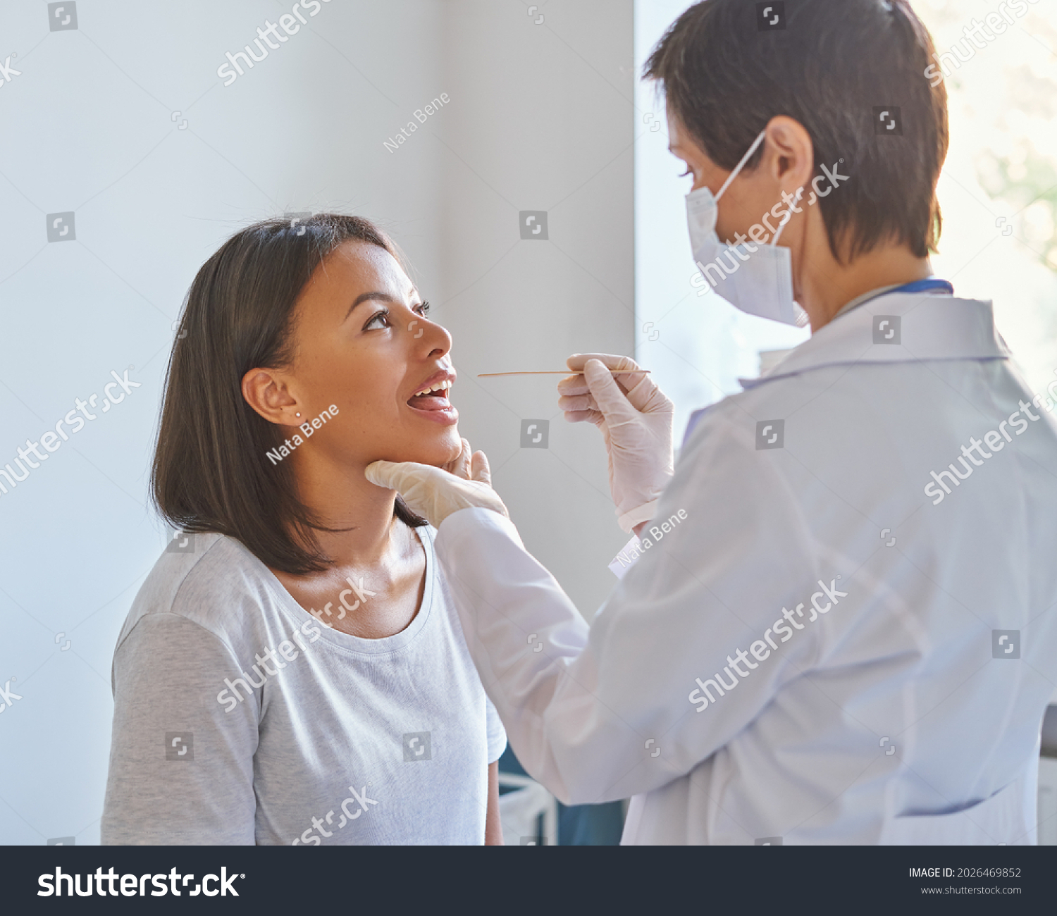 Pharyngitis and laryngitis treatment. Middle-aged woman general practitioner in face protective mask examining throat and mouth of young afro american female patient visiting ENT doctor in clinic #2026469852