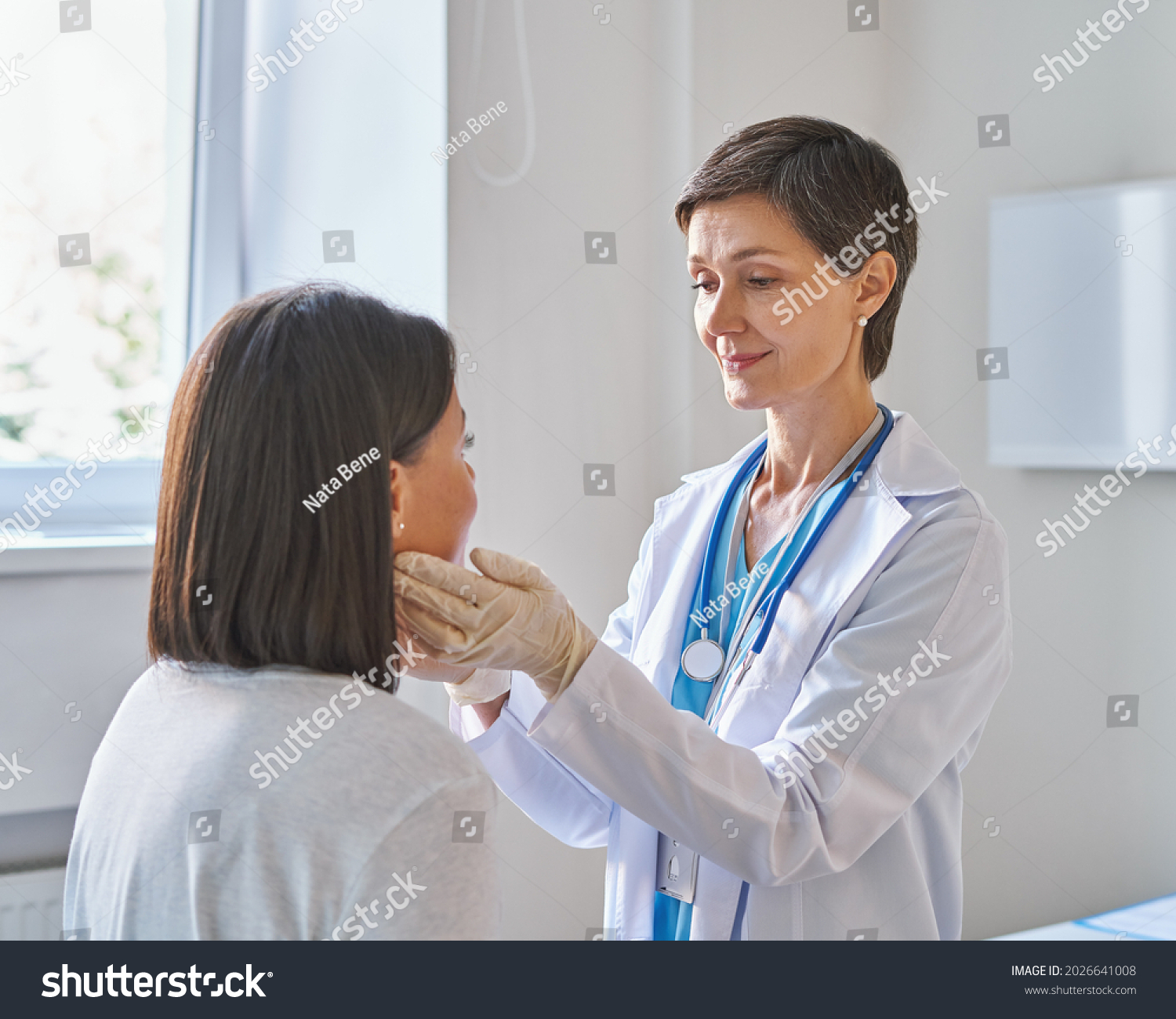 Friendly middle-aged woman doctor wearing gloves checking sore throat or thyroid glands, touching neck of young African female patient visiting clinic office. Thyroid cancer prevention concept #2026641008