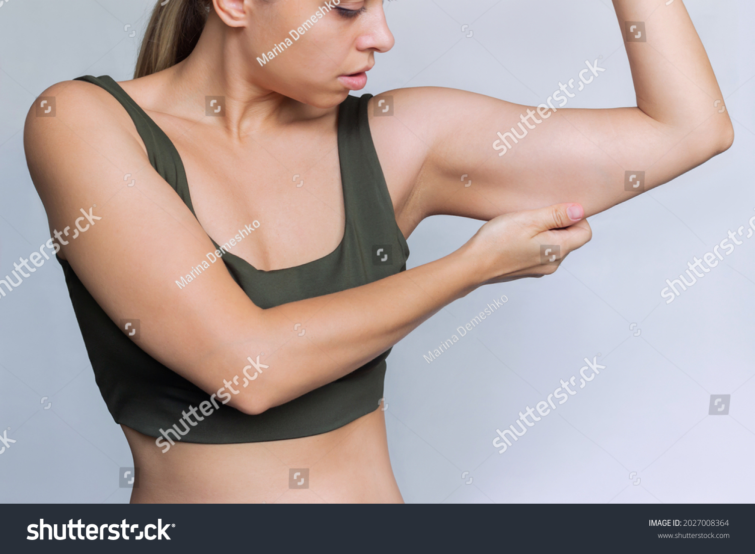 A young caucasian blonde woman  grabbing skin on her upper arm with excess fat isolated on a white background. Pinching the loose and saggy muscles. Overweight concept #2027008364