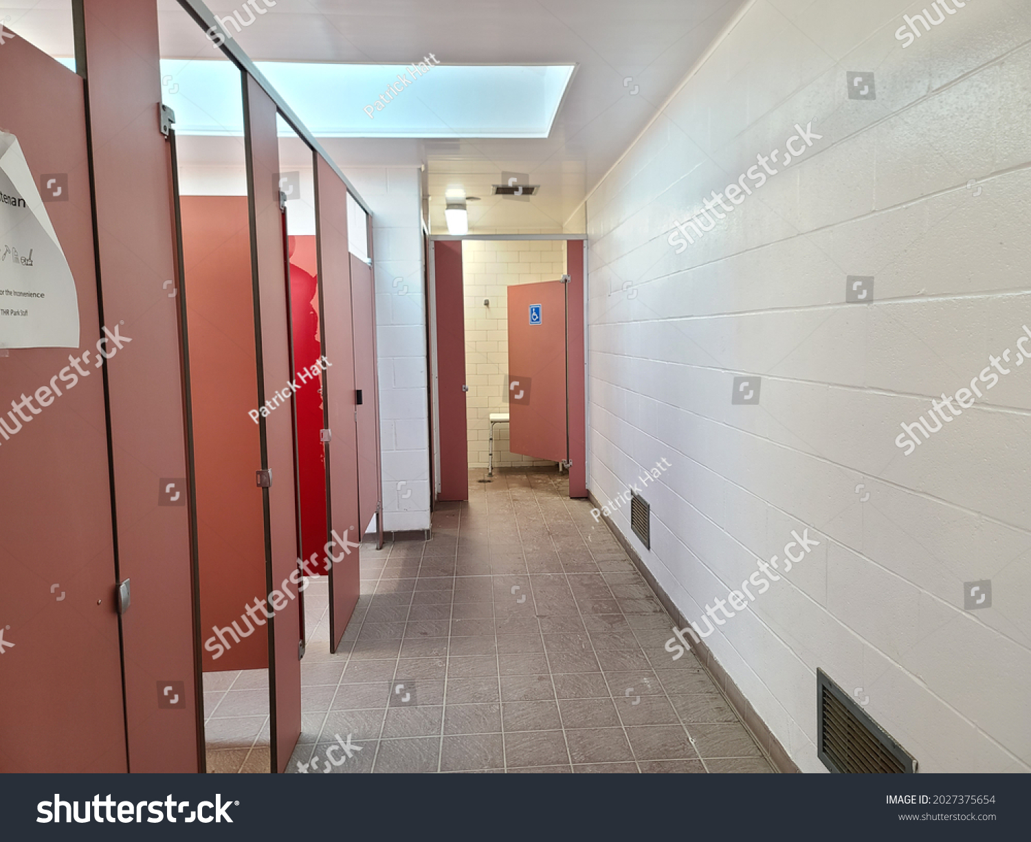 The hallway of a public bathroom. There are several salmon colored stalls in this community restroom. At the end is a larger, handicap stall with a blue handicapped symbol on it. #2027375654
