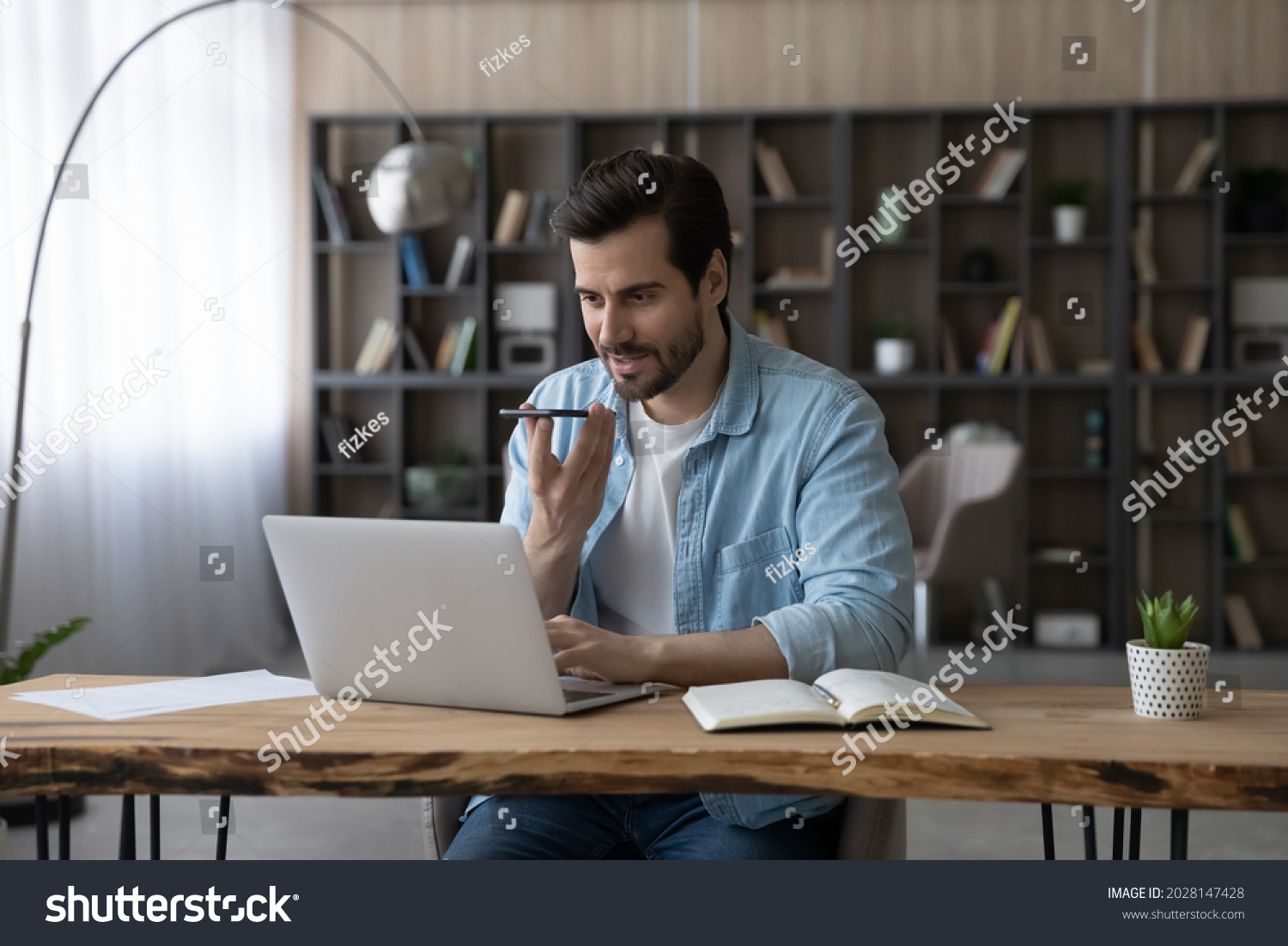 Smart skilled happy young businessman sitting at table with computer in modern home office, recording audio message for client on smartphone or holding voicemail distant conversation with colleagues. #2028147428