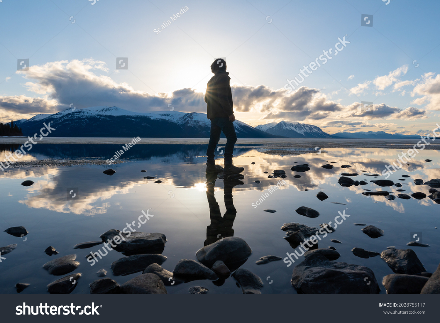 Silhouette of a man standing on a rocky lakeshore in northern Canada with reflection in calm lake below, dramatic clouds and blue sky.  #2028755117