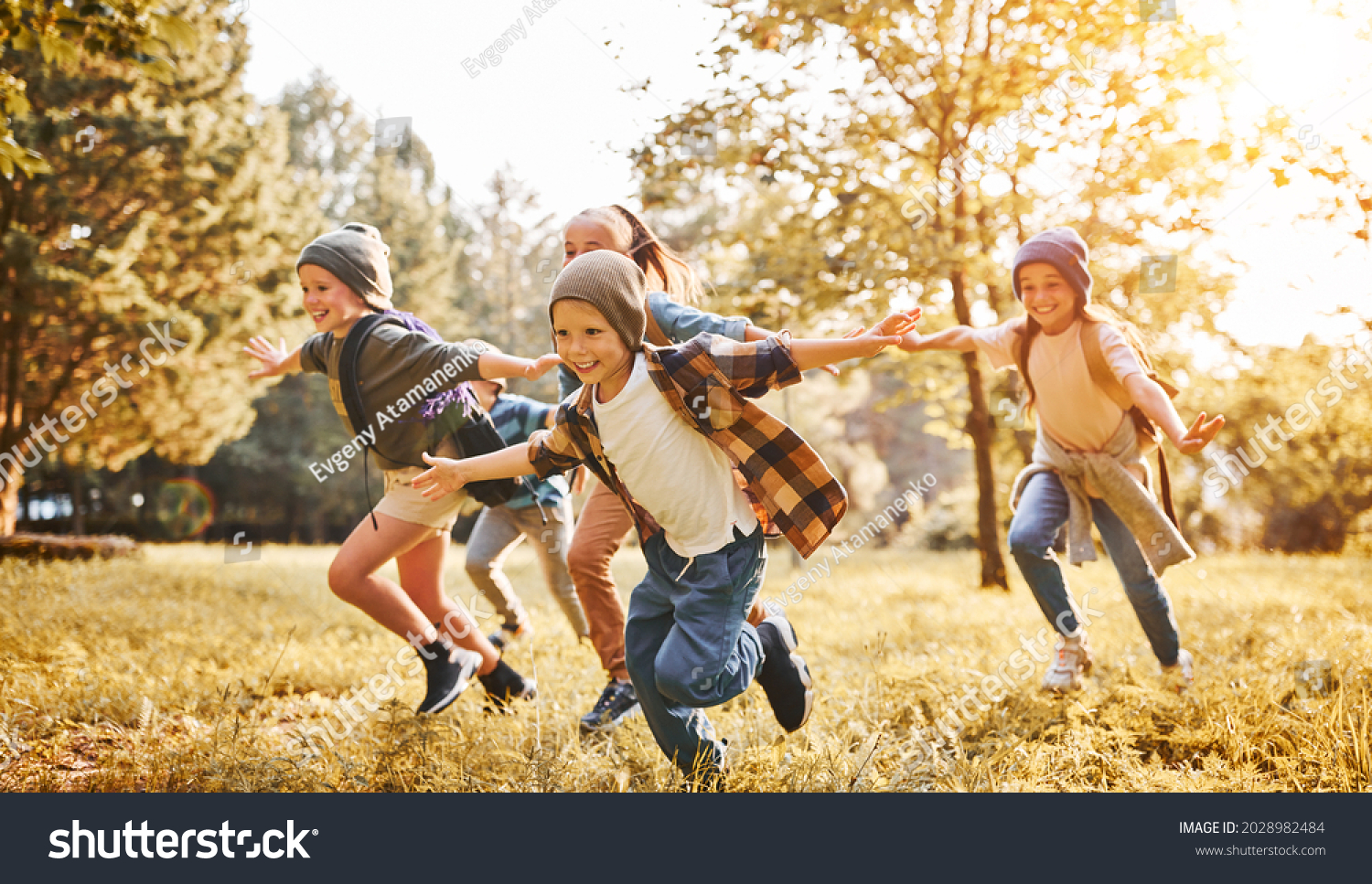 Group of happy joyful school kids with backpacks running with outstretched arms in forest on sunny spring day, excited children scouts boys and girls having fun during camping activity in nature #2028982484