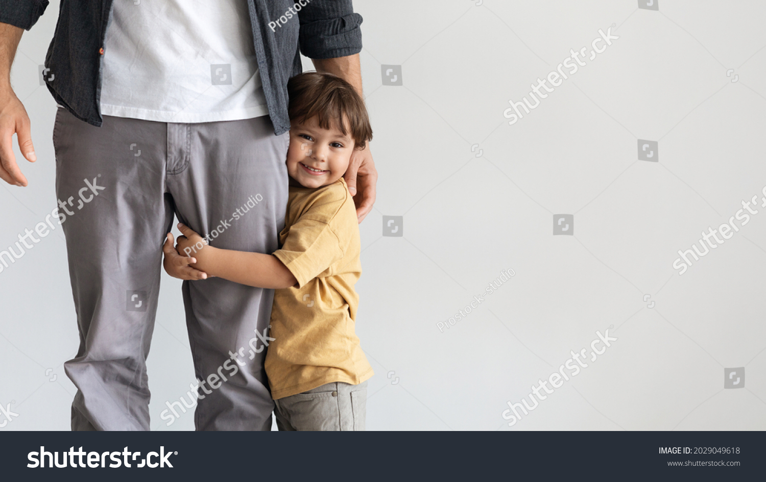 Father and son connection. Happy little boy embracing leg of his unrecognizable daddy, smiling to camera, feeling loved and safe, posing over grey wall background with empty space, panorama #2029049618
