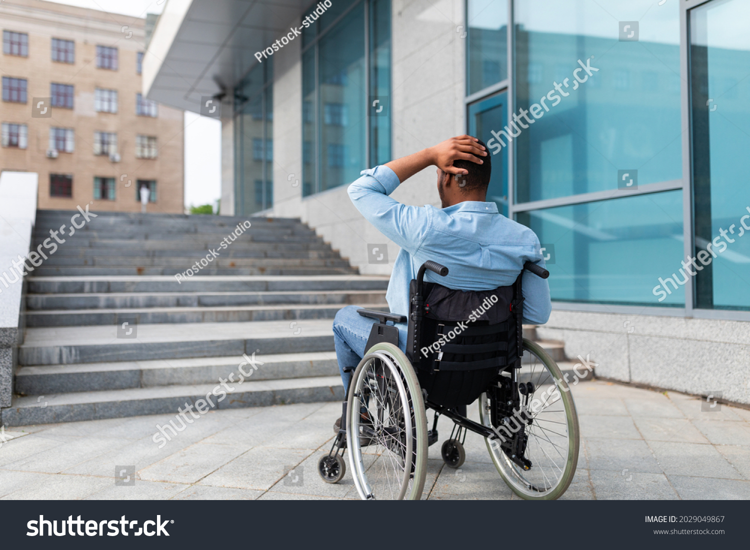 Problems with accessible environment for disabled people. Irritated impaired black man in wheelchair having no possibility to enter building without ramp, outdoors. Empty space #2029049867