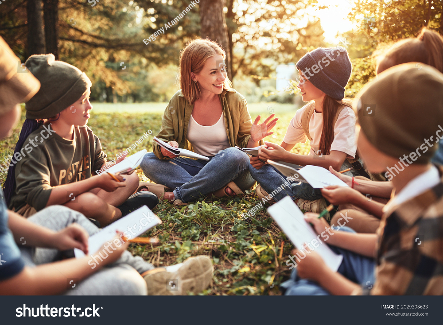 School activities in nature. Group of curious children and young female teacher sitting together on green grass in forest and learning about environment, boys and girls during ecology lesson in wood #2029398623