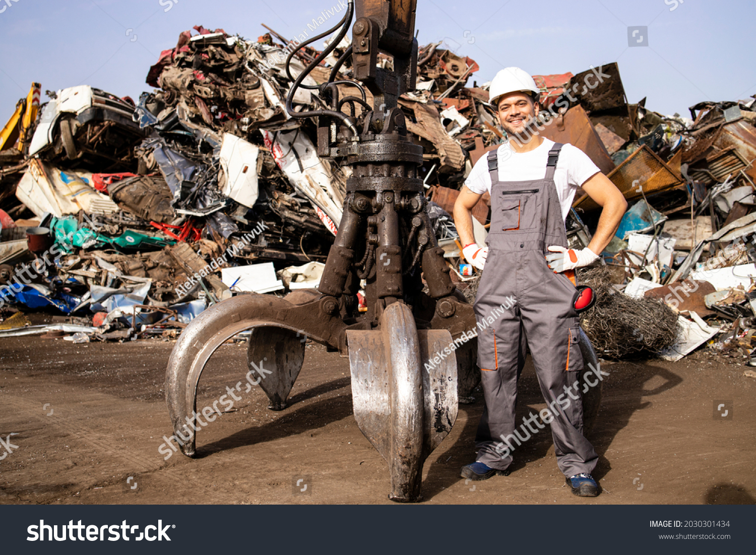 Portrait of worker standing by hydraulic industrial machine with claw attachment used for lifting scrap metal parts in junk yard. #2030301434