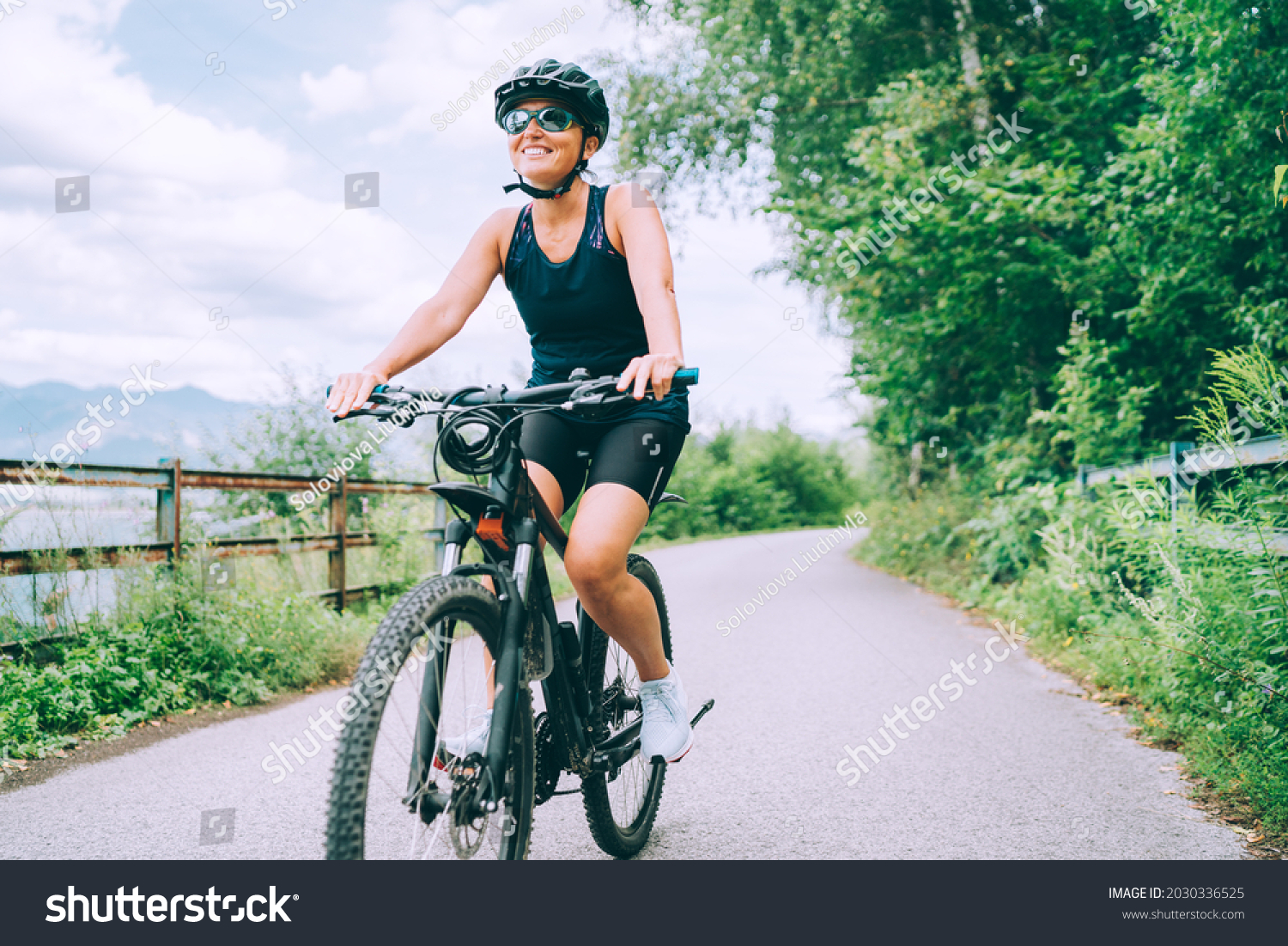 Portrait of a happy smiling woman dressed in cycling clothes, helmet and sunglasses riding a bicycle on the asphalt out-of-town bicycle path. Active sporty people concept image. #2030336525