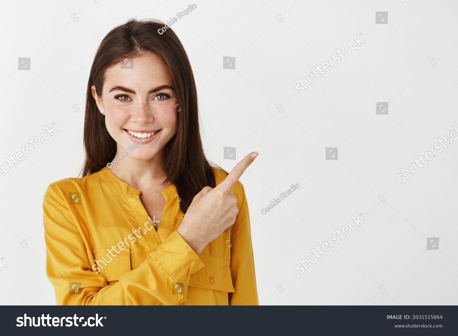Close-up of brunette female model showing promo offer, pointing finger right and smiling at camera, white background #2031515864